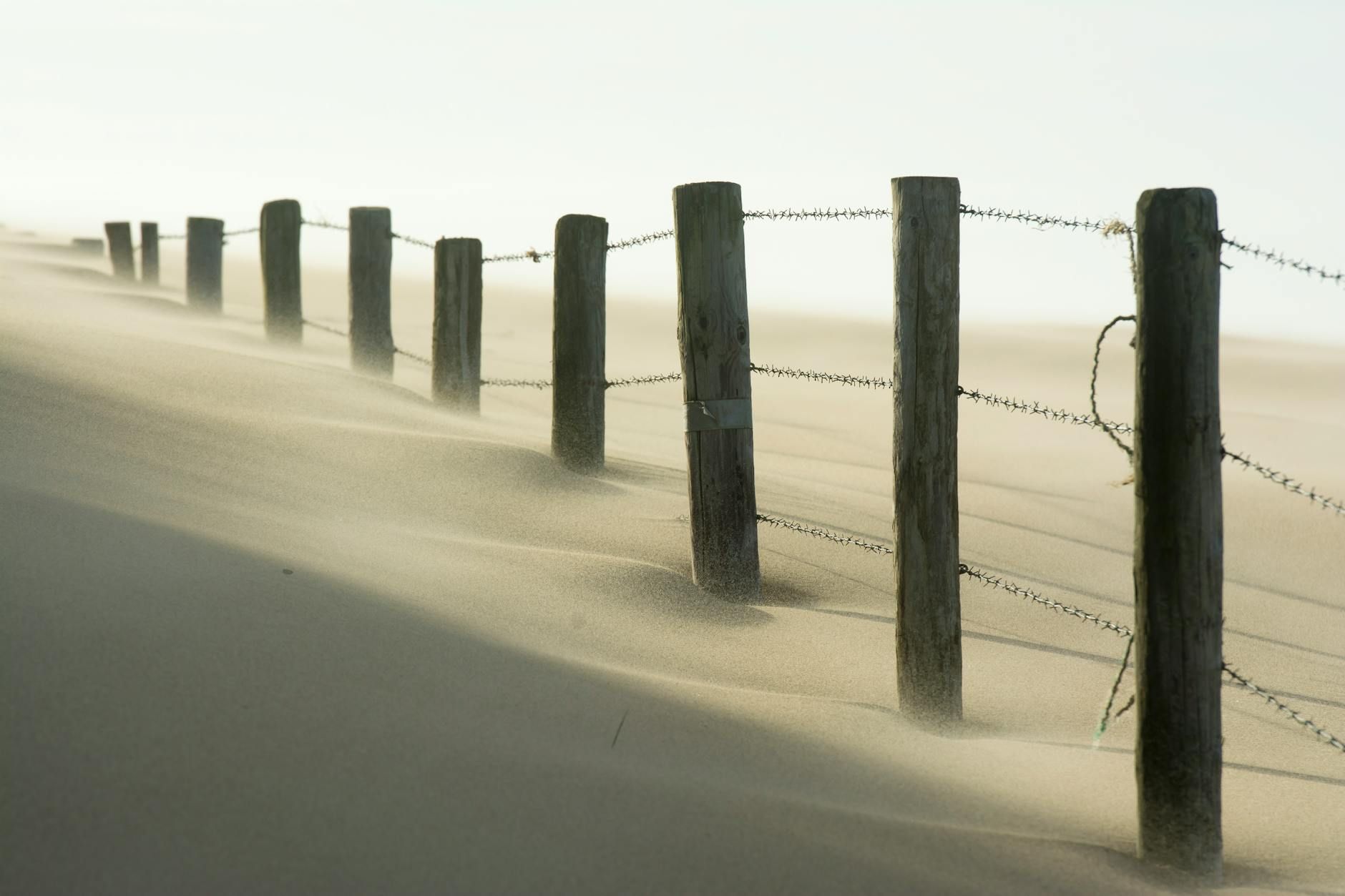 A scenic view of a barbed wire fence stretching across sandy dunes, with wind blowing sand.