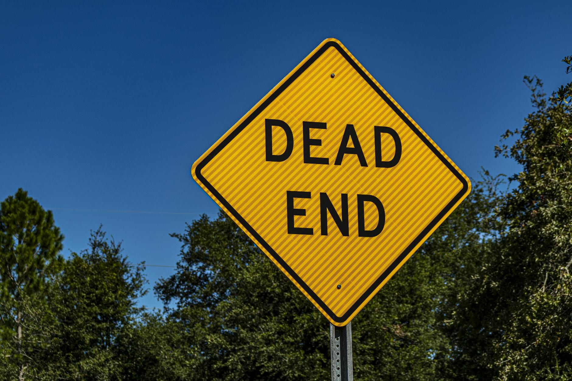 Close-up of a yellow 'Dead End' sign against a blue sky and trees, symbolizing caution and warning.
